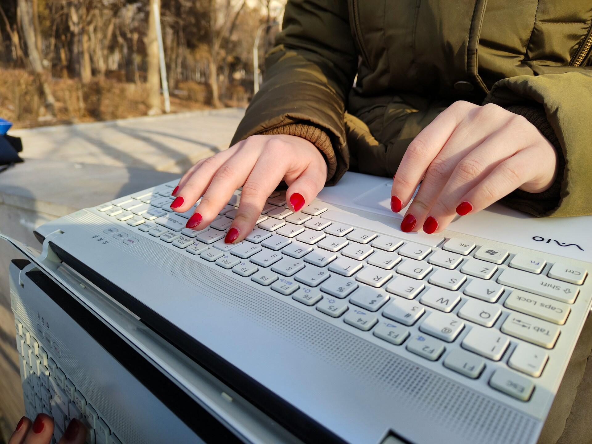 A close-up of hands with red nail polish typing on a laptop keyboard outdoors, sunlight casting shadows on the surface.