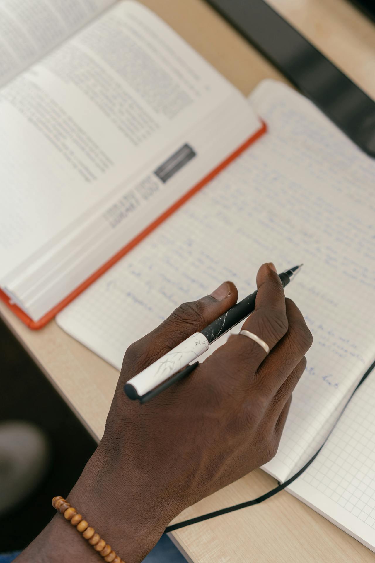 A close-up of a hand holding a black and white pen, writing in a notebook next to an open book on a desk.