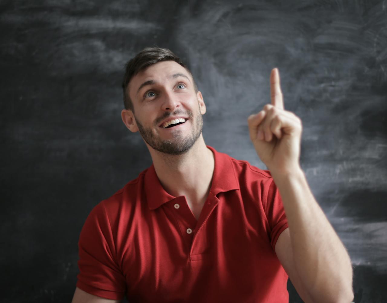 A person wearing a red polo shirt raises one finger, suggesting a point or idea, against a dark chalkboard background.