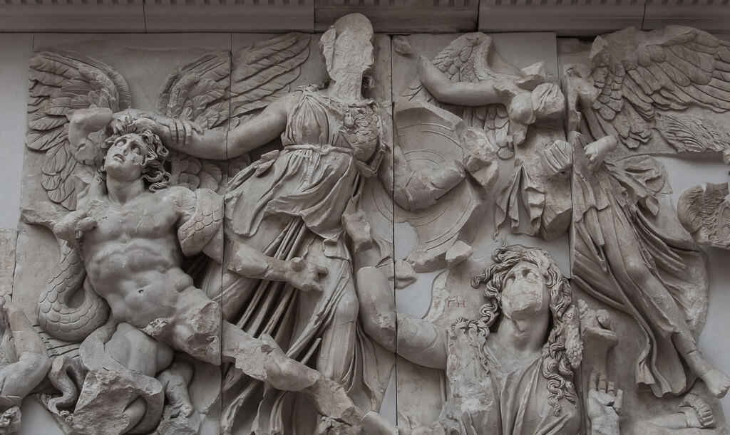 Ancient stone relief depicting mythological figures in dynamic poses. A winged male figure struggles on the left, while central figures engage in battle.