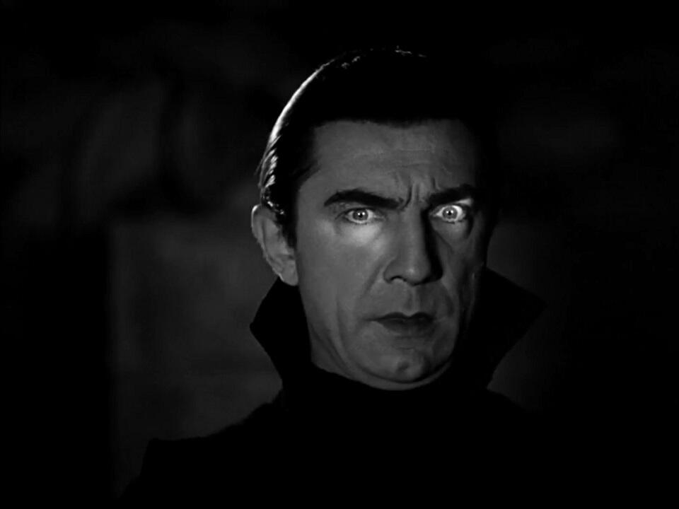 Black-and-white still of Bela Lugosi in costume as Count Dracula, wearing a dark cape and formal evening wear, with his hand raised in a dramatic, sinister gesture (1931 film).