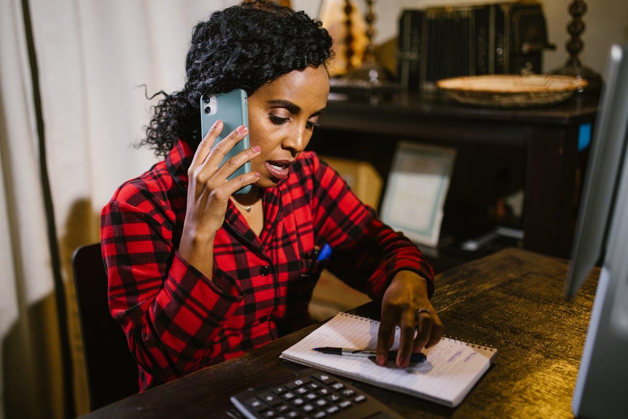A person in a red plaid shirt speaks on a smartphone while taking notes in a notepad at a wooden desk, with a computer and decor in the background.