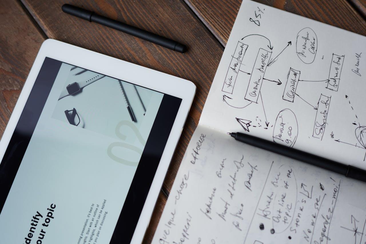 Close-up of a tablet displaying design content, alongside a notebook filled with handwritten notes and diagrams, and a pen.