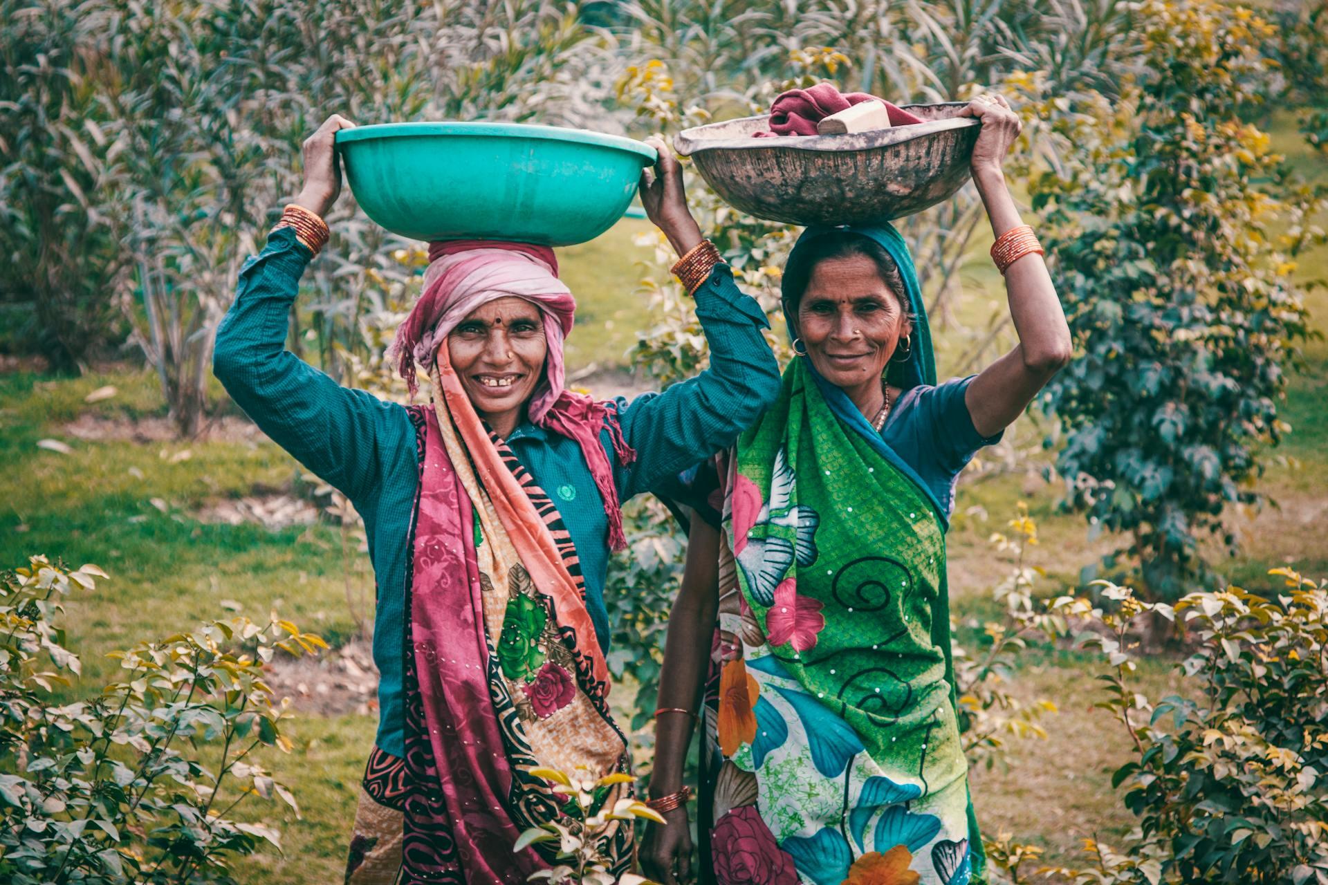 Two Indian women dressed in colourful clothes holding bowls above their heads 
