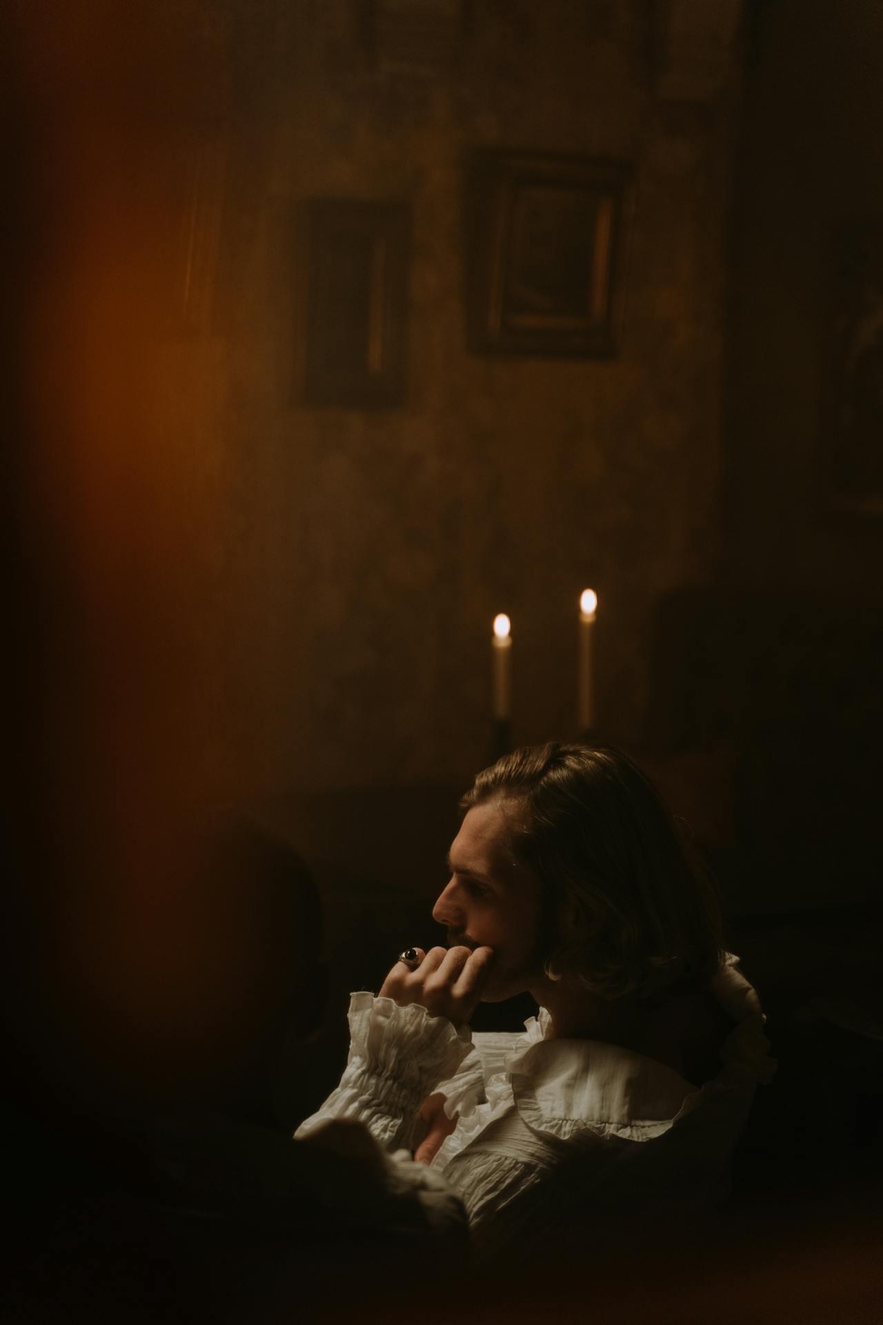 A dimly lit room with soft candlelight, featuring a person in a ruffled shirt engaged in contemplation, surrounded by antique decor.