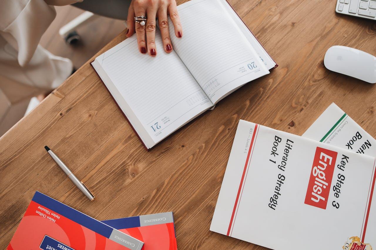 A hand with red nails rests on an open planner, surrounded by books and a pen on a wooden desk, suggesting study or planning.