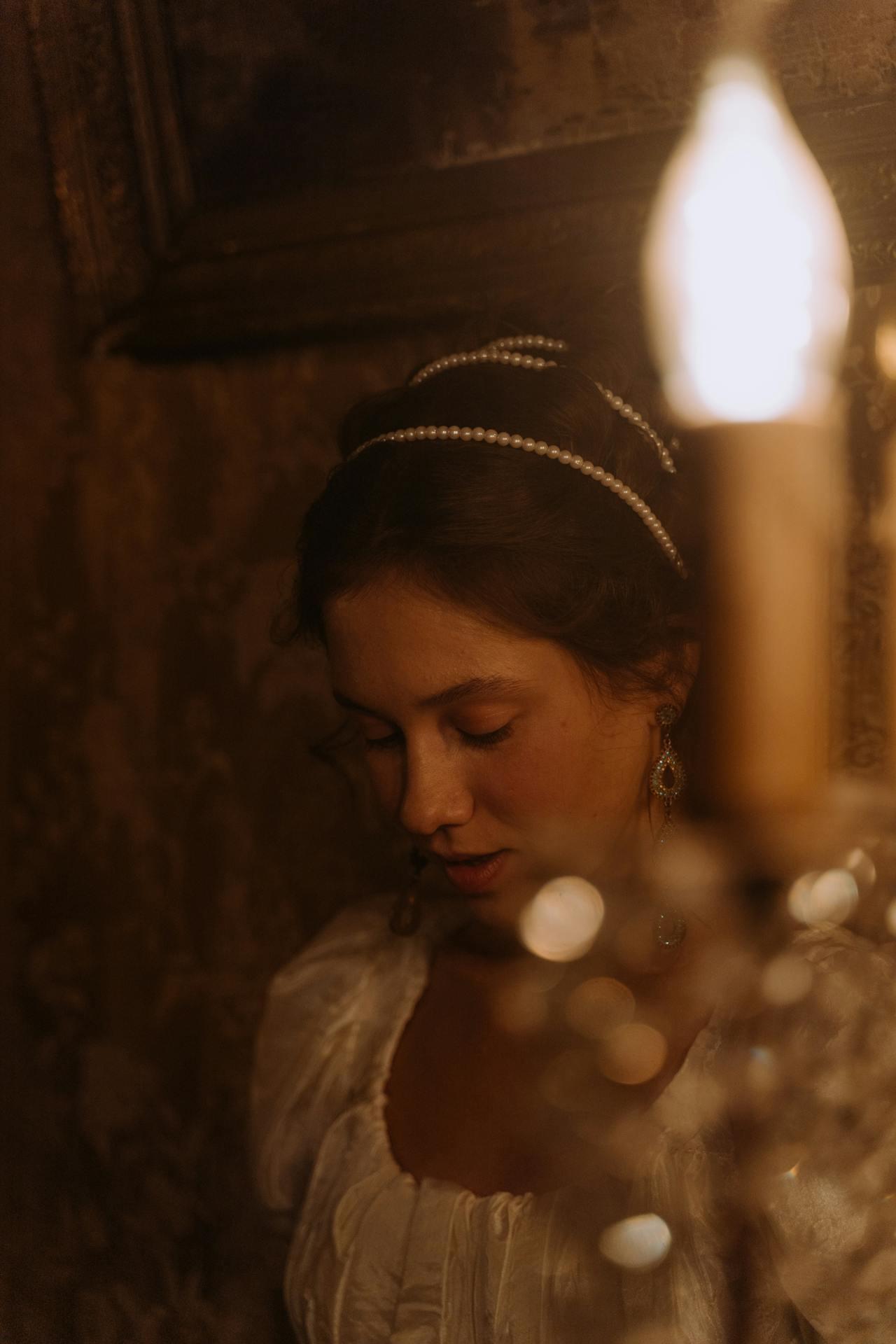 A woman in a vintage white dress and pearl accessories stands near a flickering candle, surrounded by dim, ornate wallpaper.