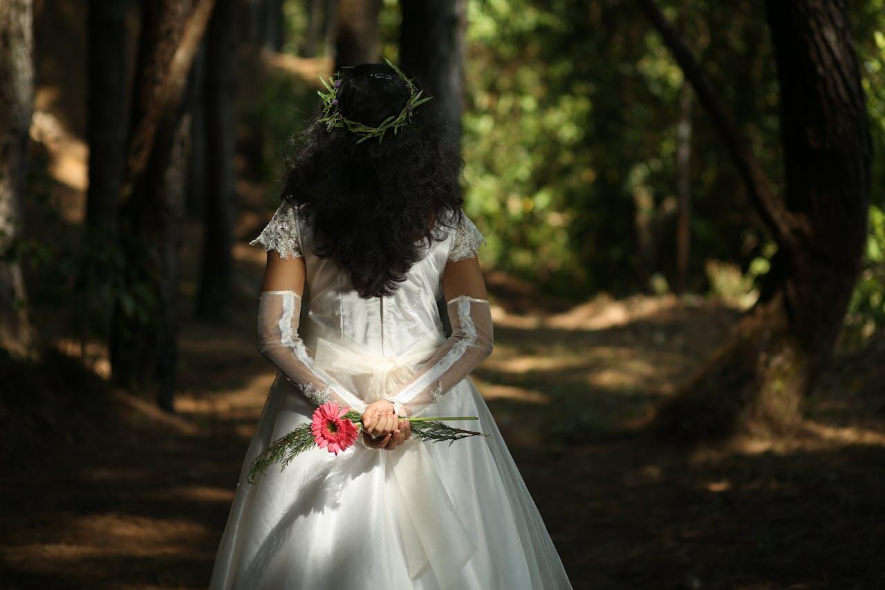 A bride in a flowing white gown and floral crown stands in a sunlit forest, holding pink flowers behind her back.