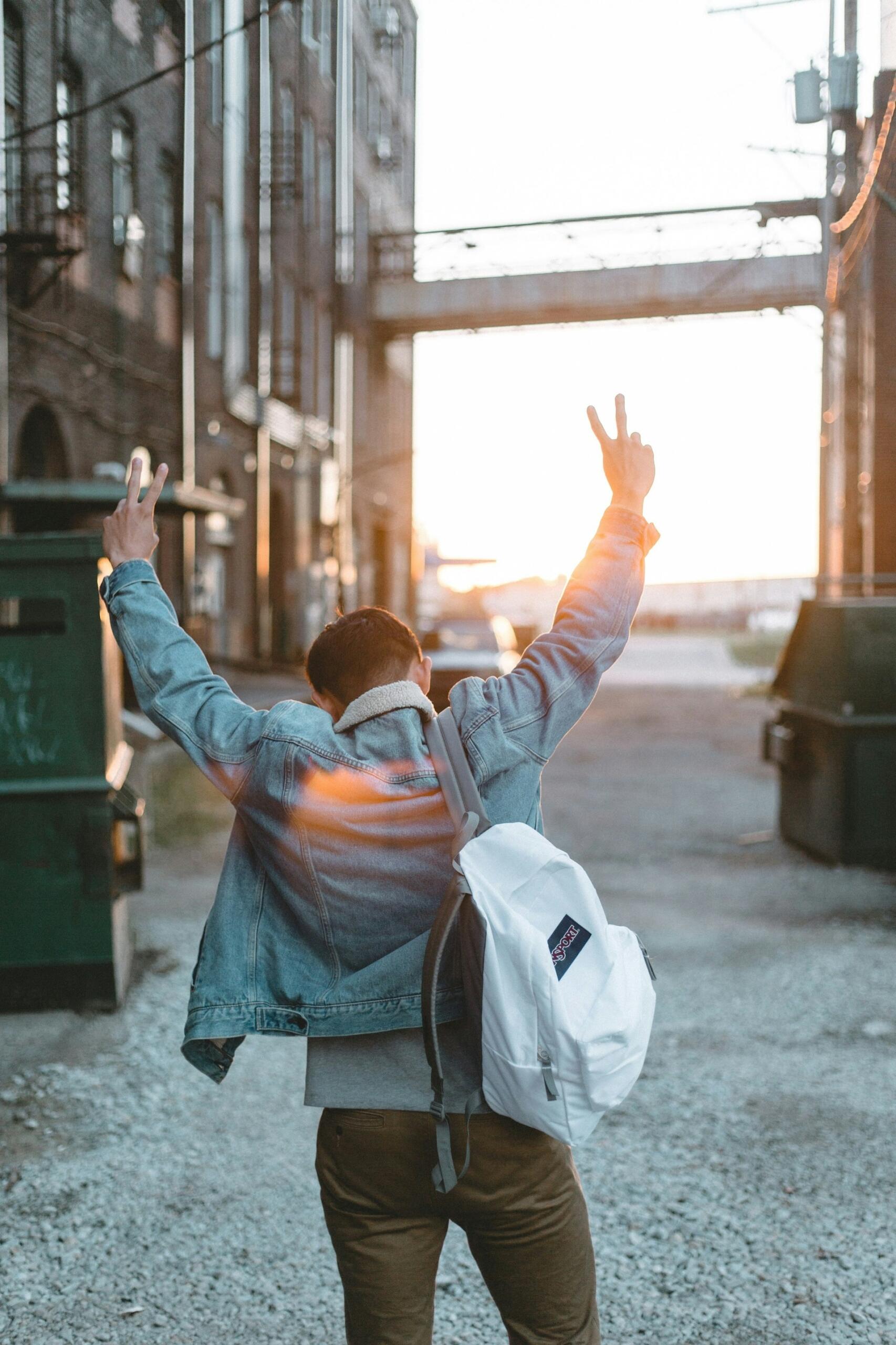 A person in a jeans jacket with a white backpack holds their arms up in a victory pose. 
