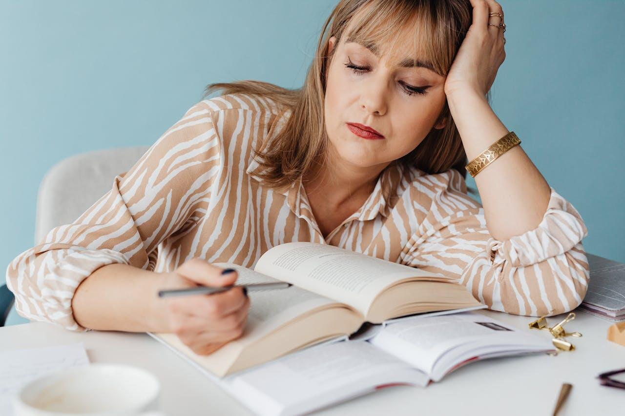 A person in a striped blouse rests their head on their hand while studying a large book at a desk, surrounded by notes and a cup.