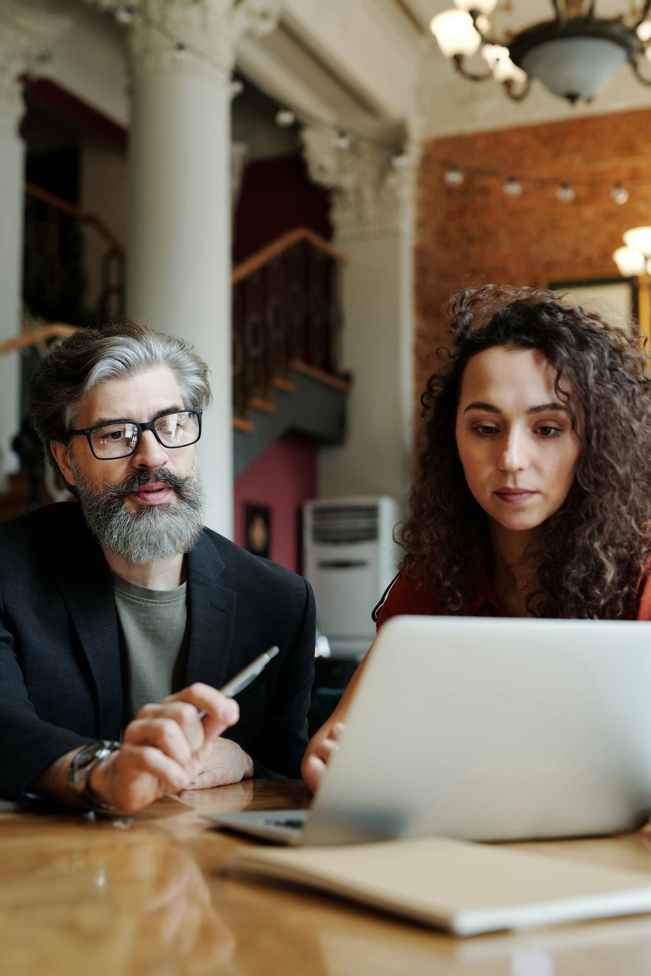 Two people engage in a discussion over a laptop on a wooden table, surrounded by an elegant interior.