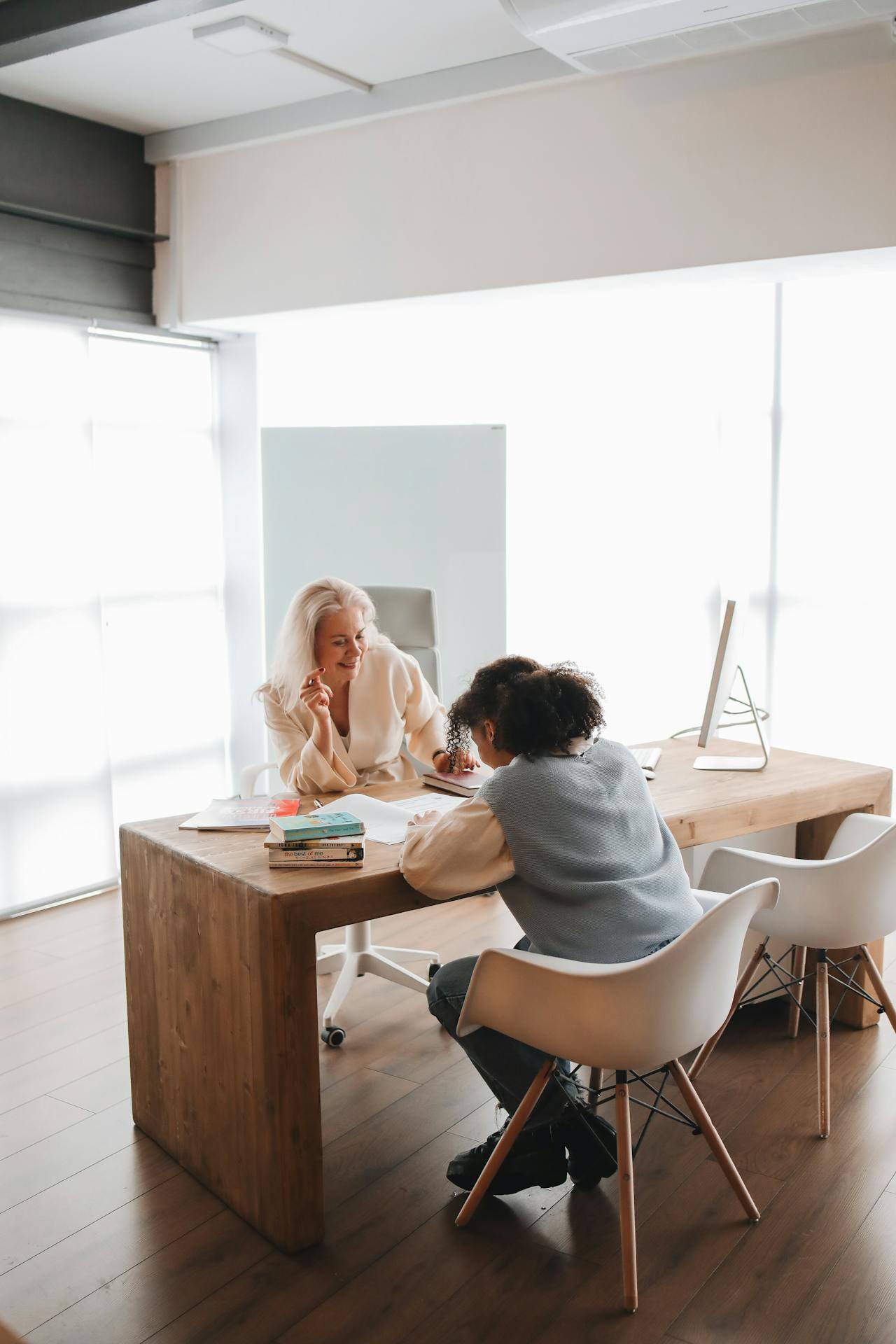 Two people sit at a wooden table in a bright office, discussing documents with books and a computer visible nearby.