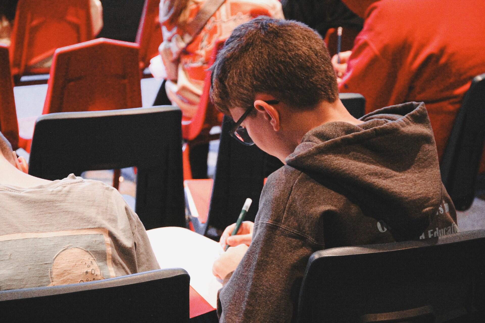 A student in a gray hoodie sits at a desk, writing notes in a notebook during a classroom session with classmates in the background.