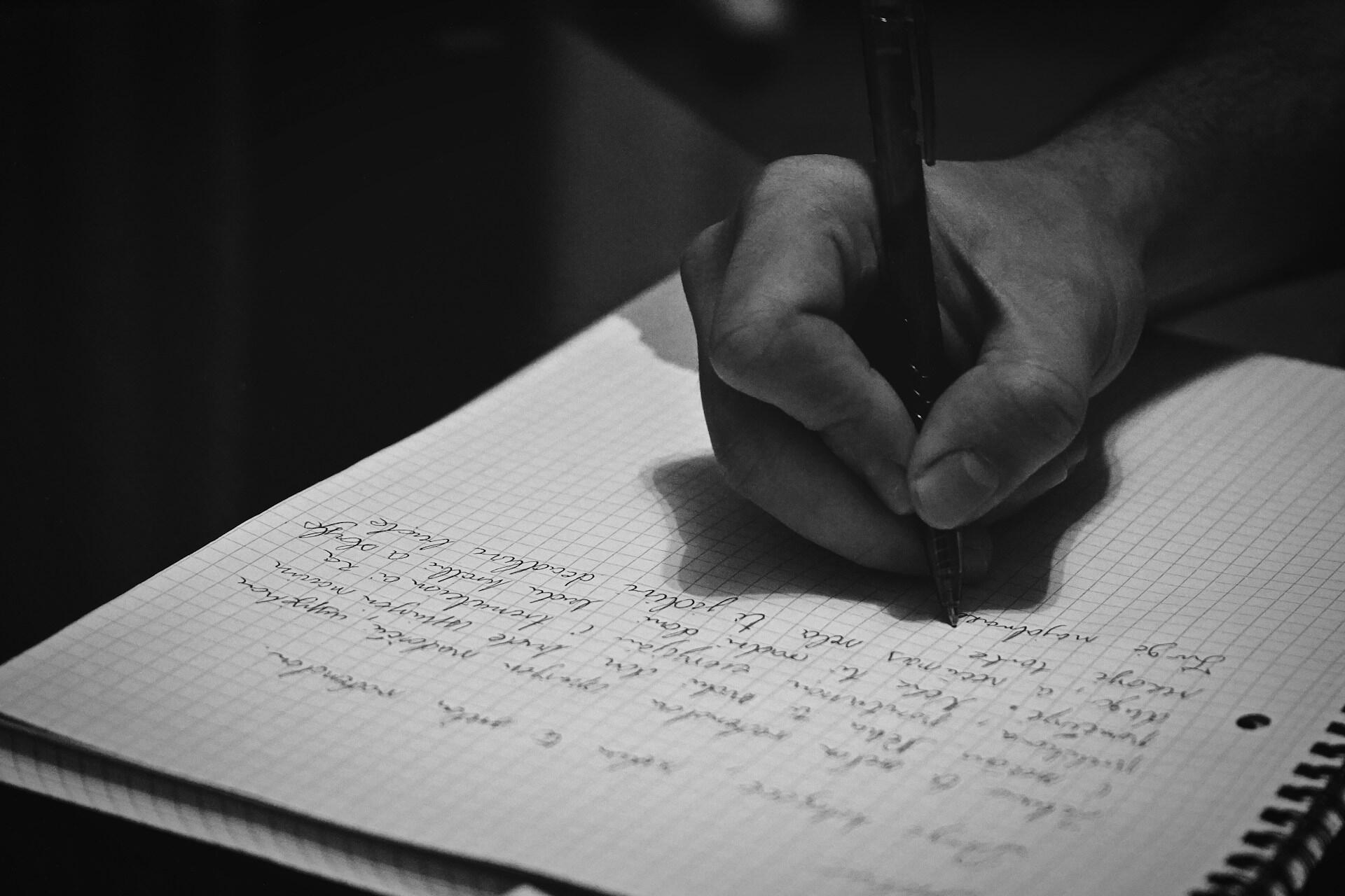 A hand holding a pen writes on a grid-patterned notebook page, filled with handwritten notes in a black and white setting.