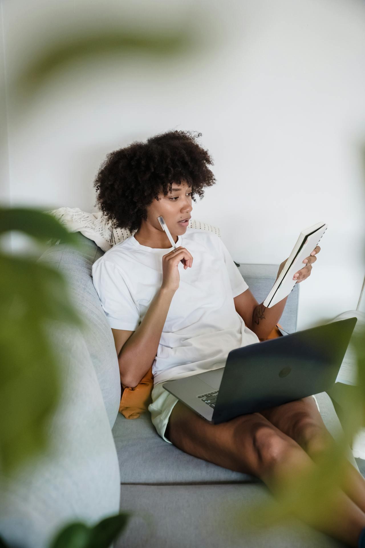A person sits comfortably on a couch, holding a tablet in one hand while a laptop rests on their lap, surrounded by greenery.