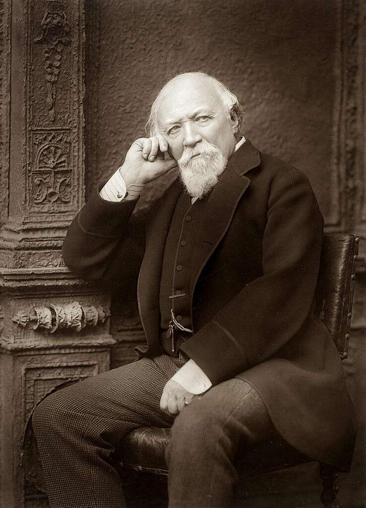 A seated elderly man in formal attire, thoughtfully resting his chin on his hand, against an ornate background.