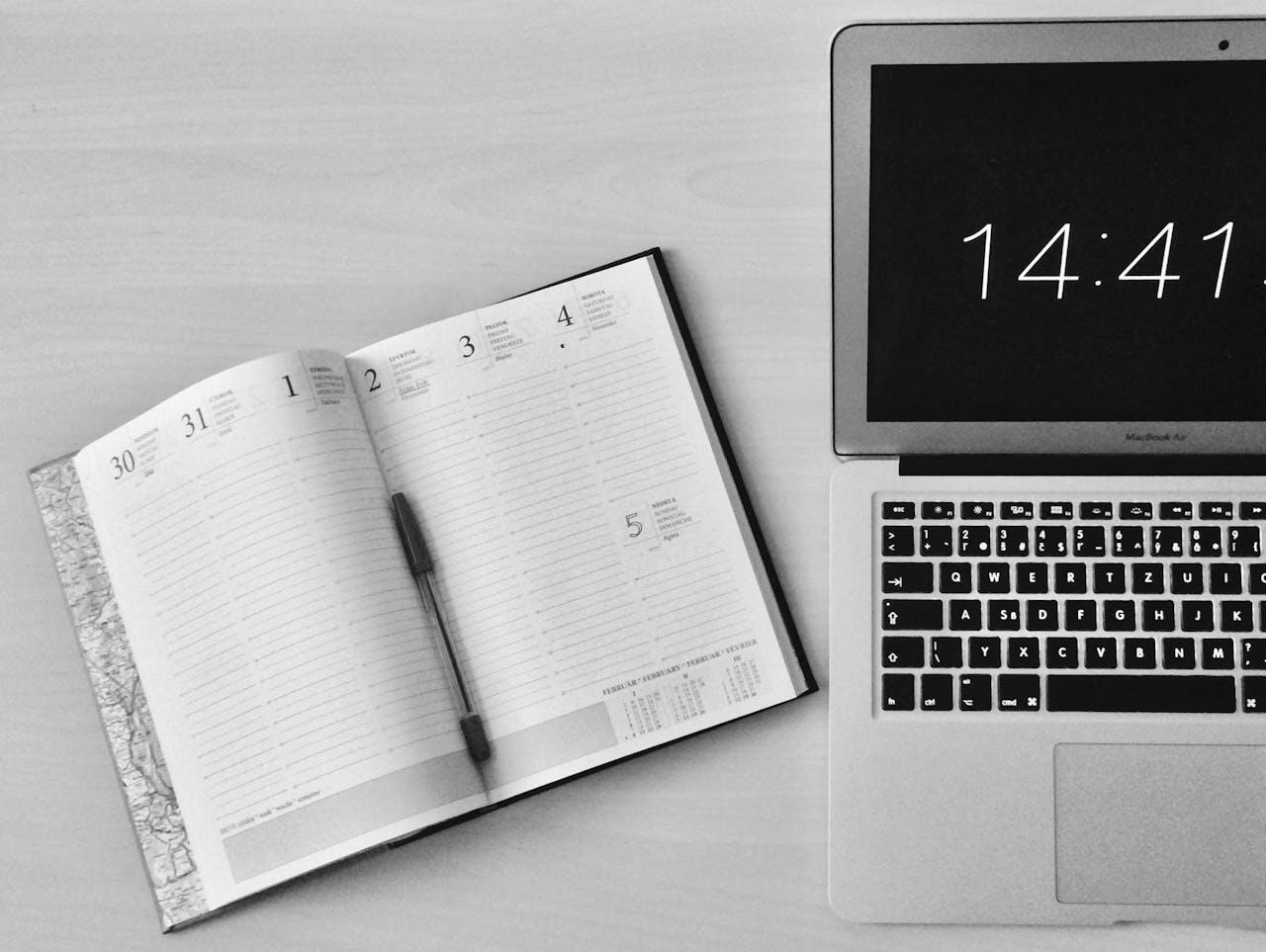 A black and white image of an open planner with a pen beside it, next to a MacBook Air displaying the time 14:41.