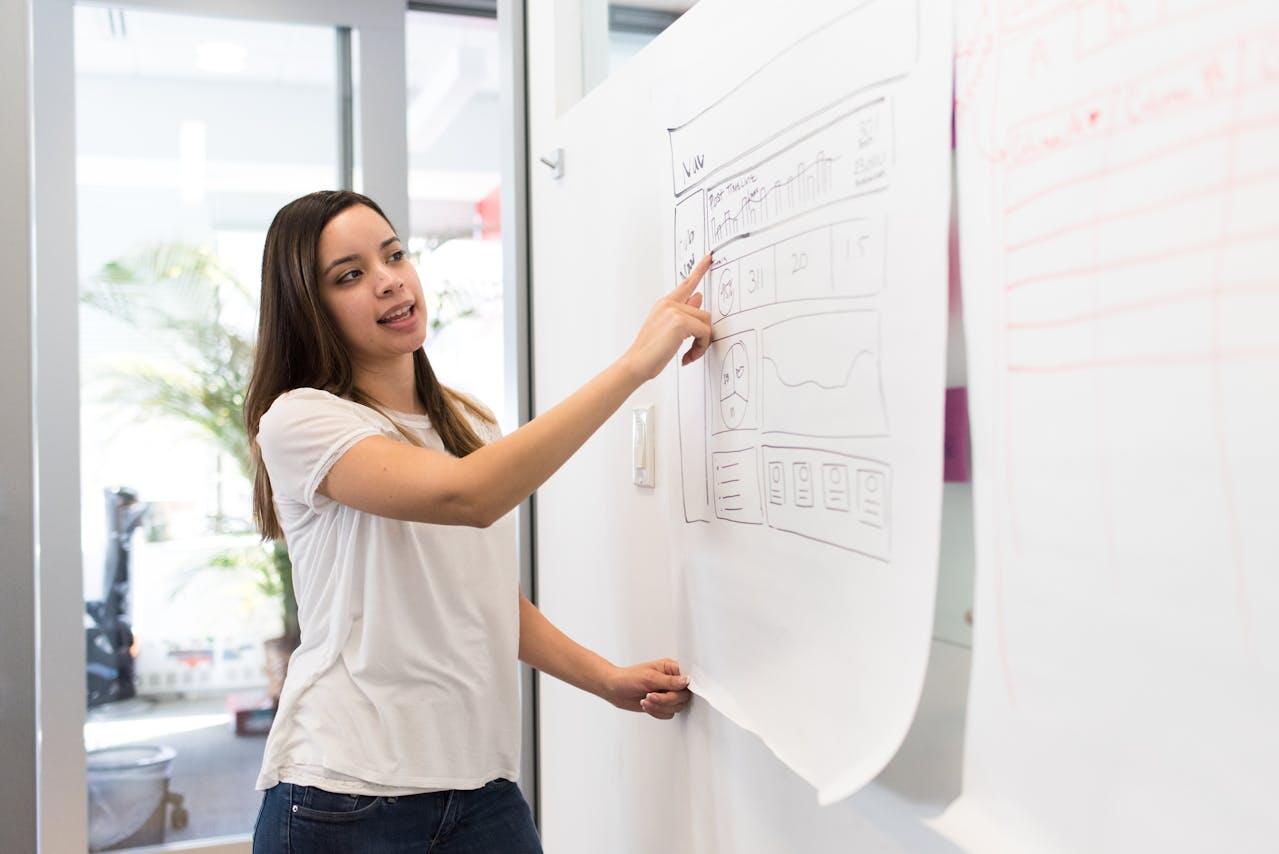 A woman in a casual white t-shirt points to a large white paper displaying sketches and charts in a bright, modern office space.
