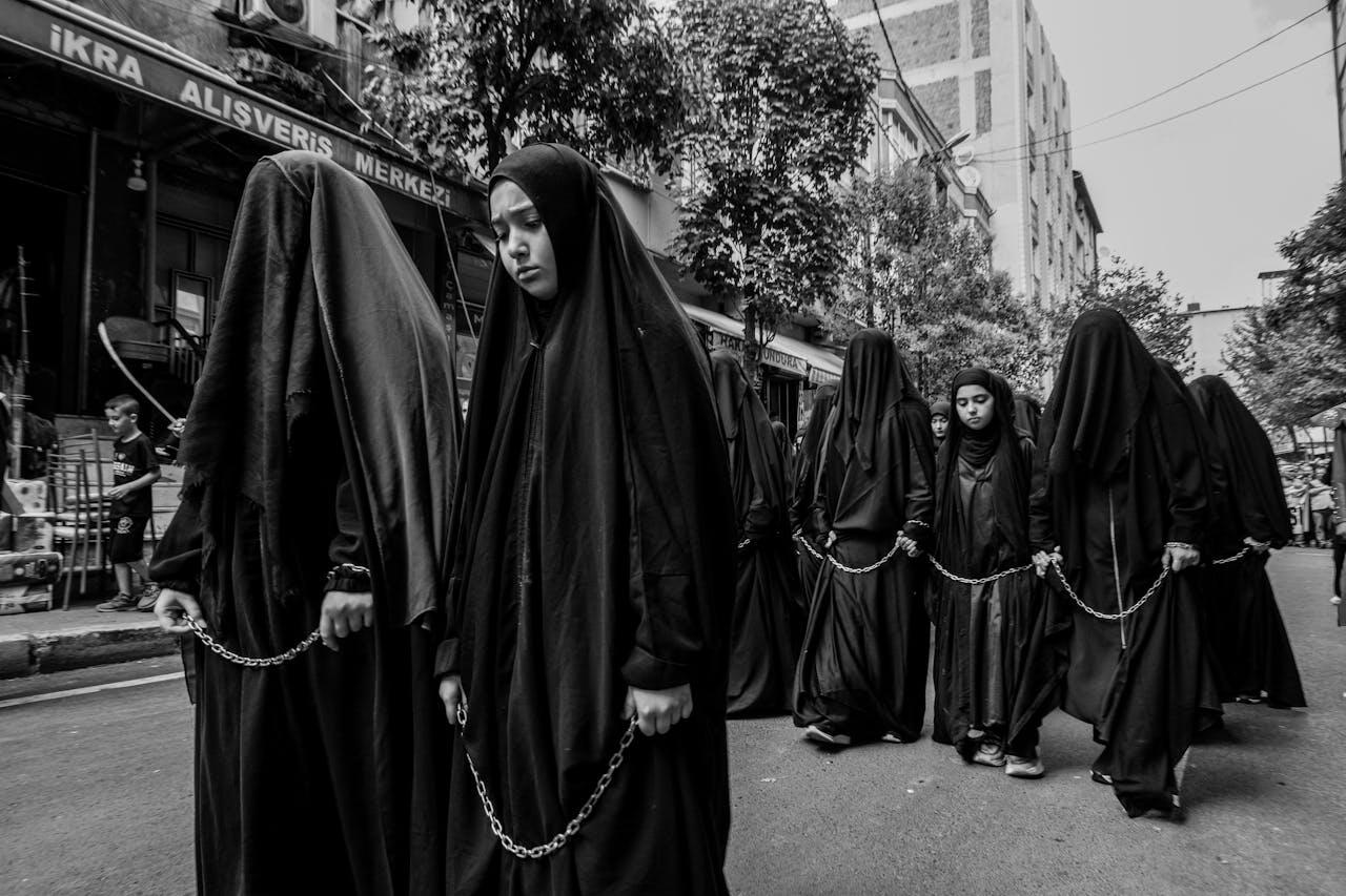 A group of individuals in long black cloaks walk in a procession down a street, linked by chains, surrounded by urban scenery.