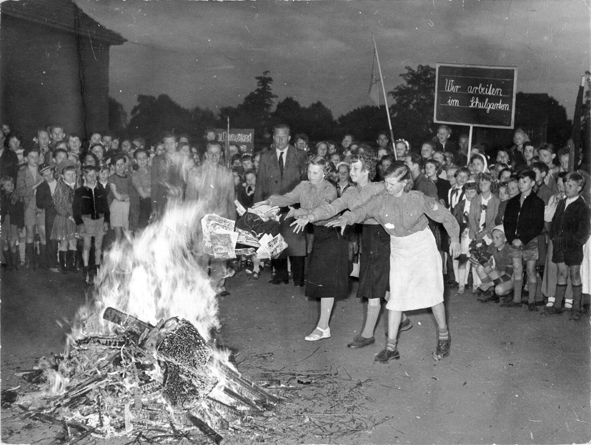black and white photo of adult women throwing books into a fire in front of a group of children