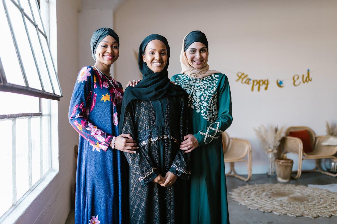 Three women in elegant attire stand together, celebrating with a "Happy Eid" banner in a cozy, well-lit room.