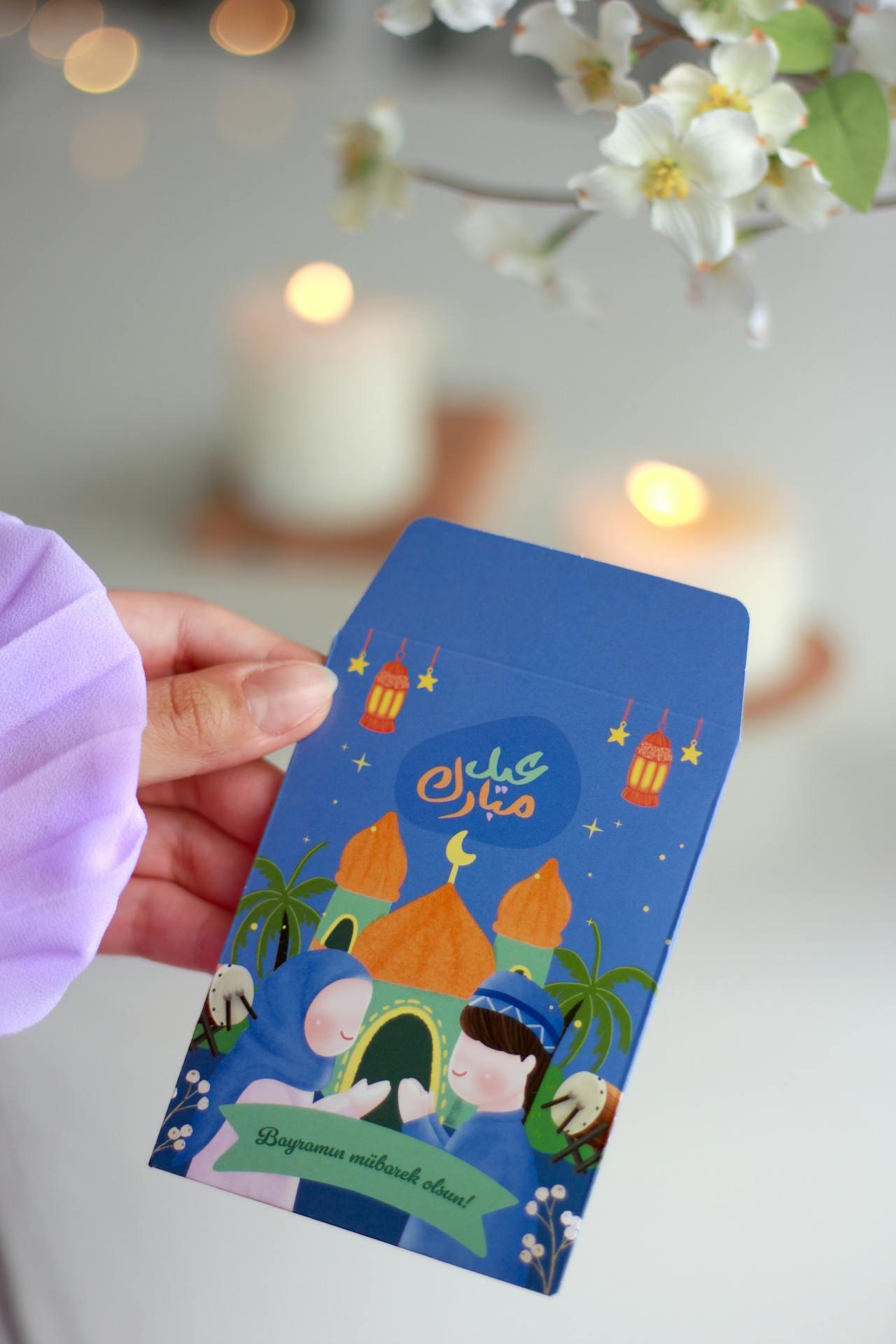 A hand holds a colorful envelope featuring a festive design with Muslim characters, mosques, and lanterns, celebrating a special occasion.