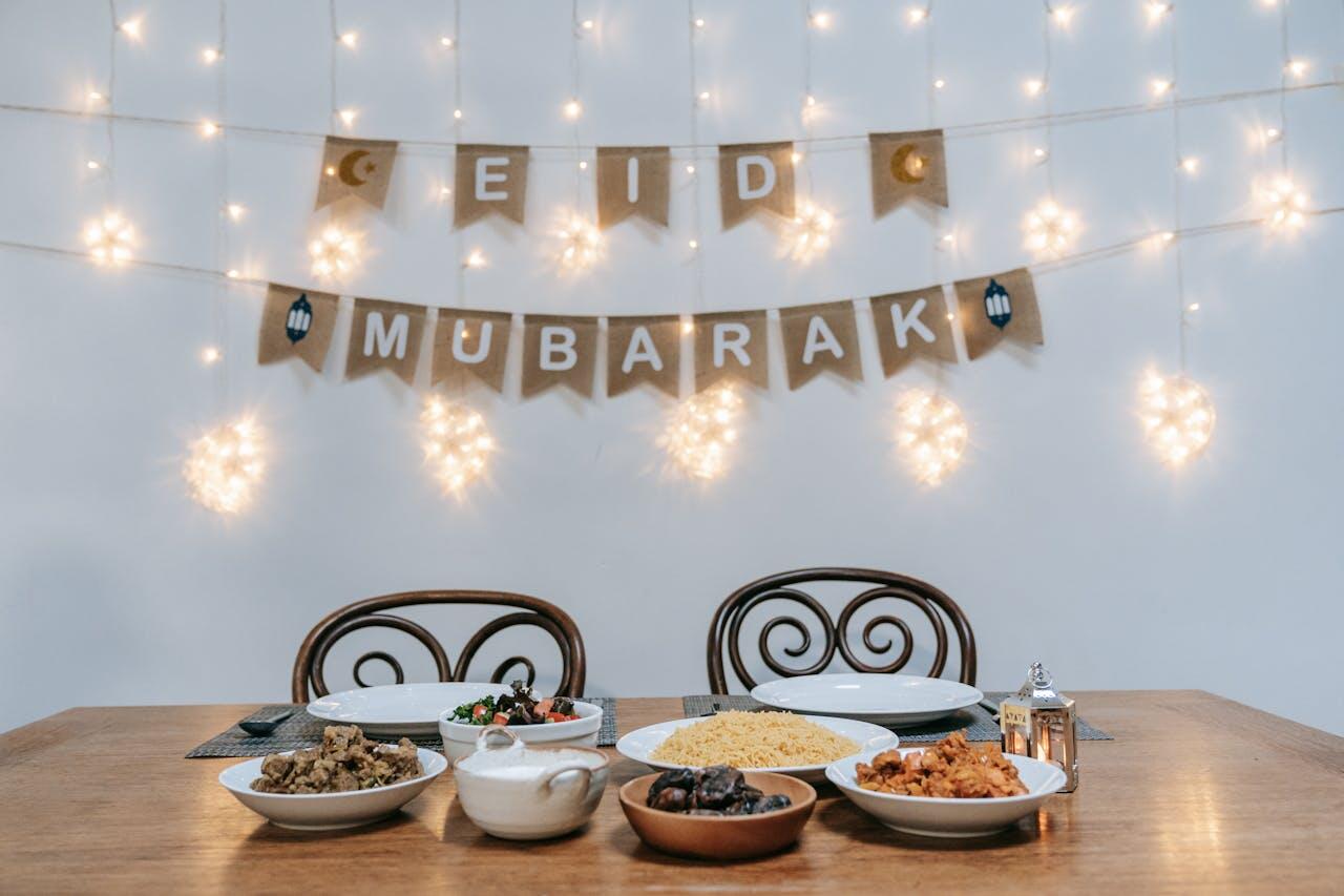 A festive table set for Eid Mubarak, featuring various dishes and desserts, with a decorative banner and twinkling lights in the background.