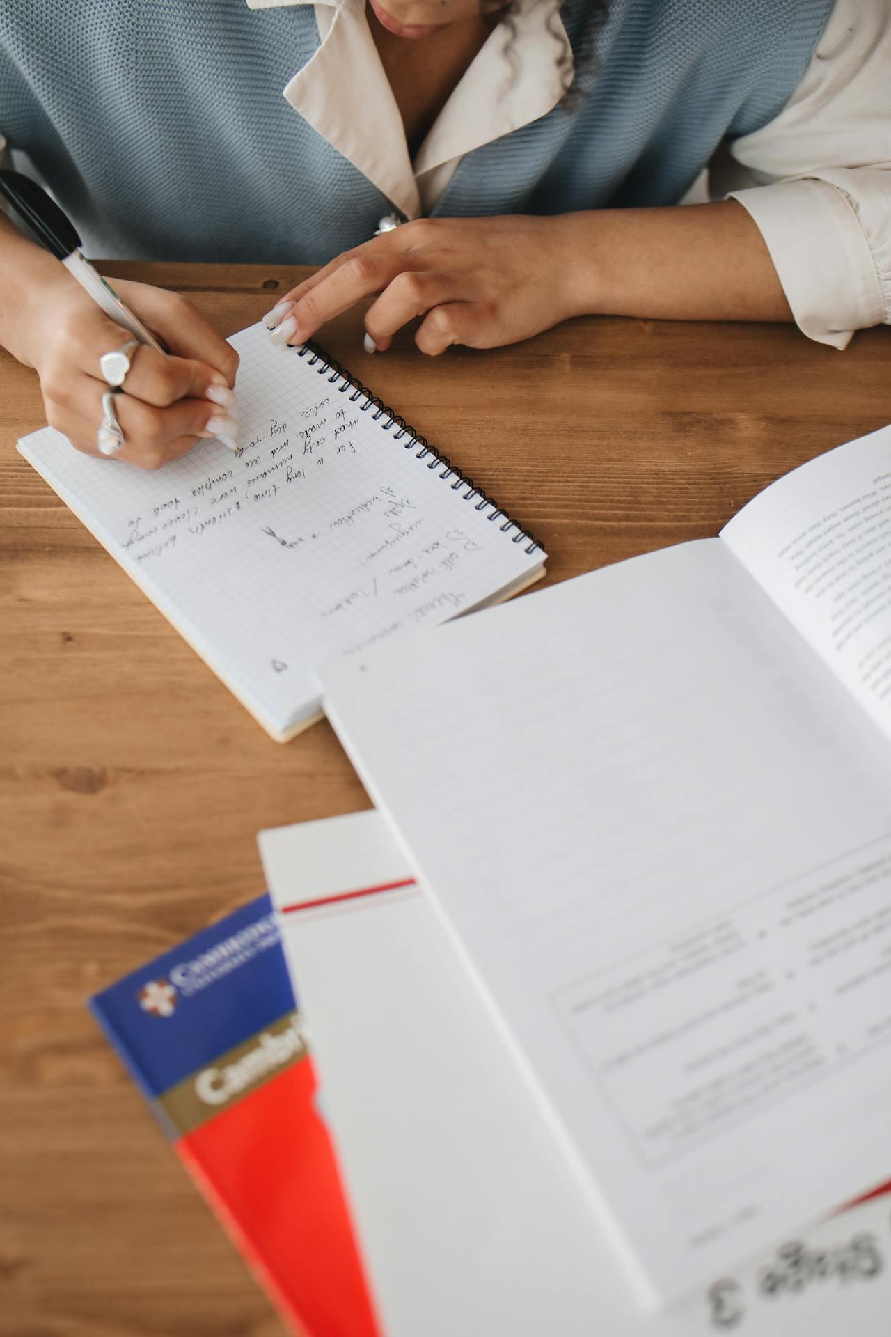 A person writes notes in a spiral notebook, surrounded by notebooks and textbooks on a wooden table.