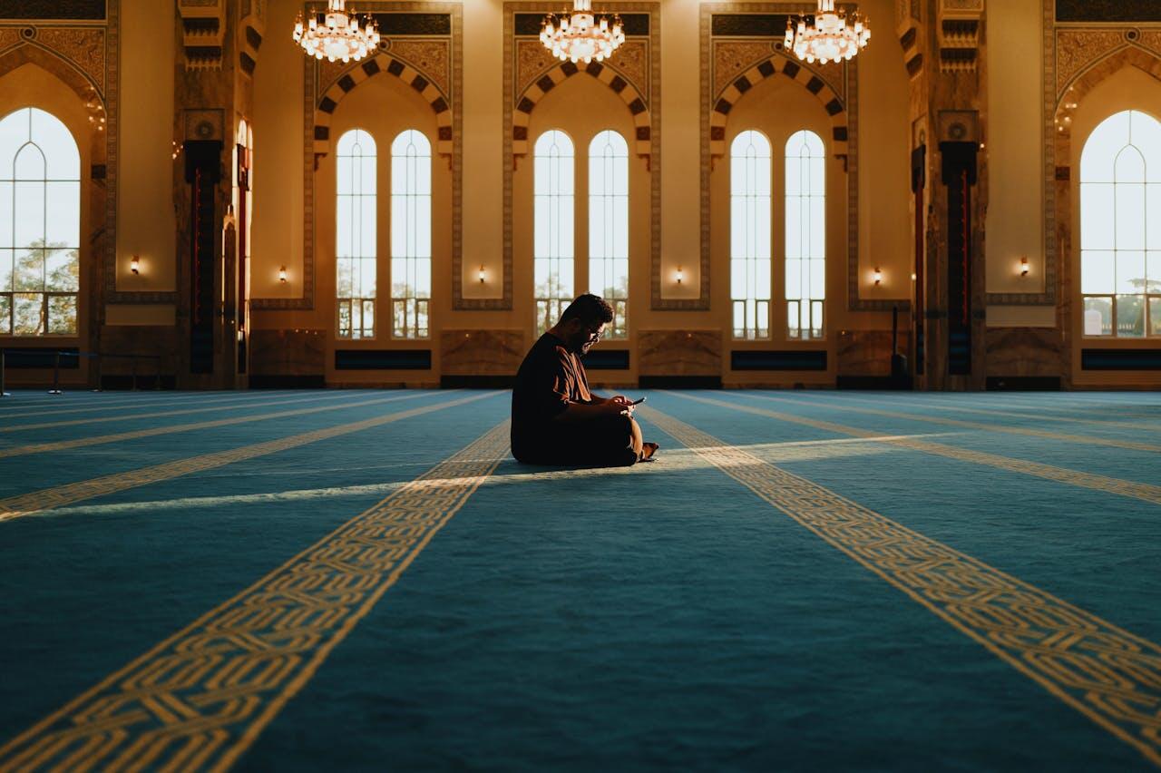 A person sits on a vibrant blue prayer rug inside a sunlit mosque, surrounded by elegant architecture and tall, arched windows.