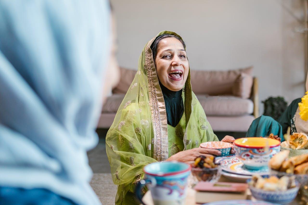 A woman in a vibrant green outfit sits at a table filled with colorful dishes, engaging in conversation with another person.