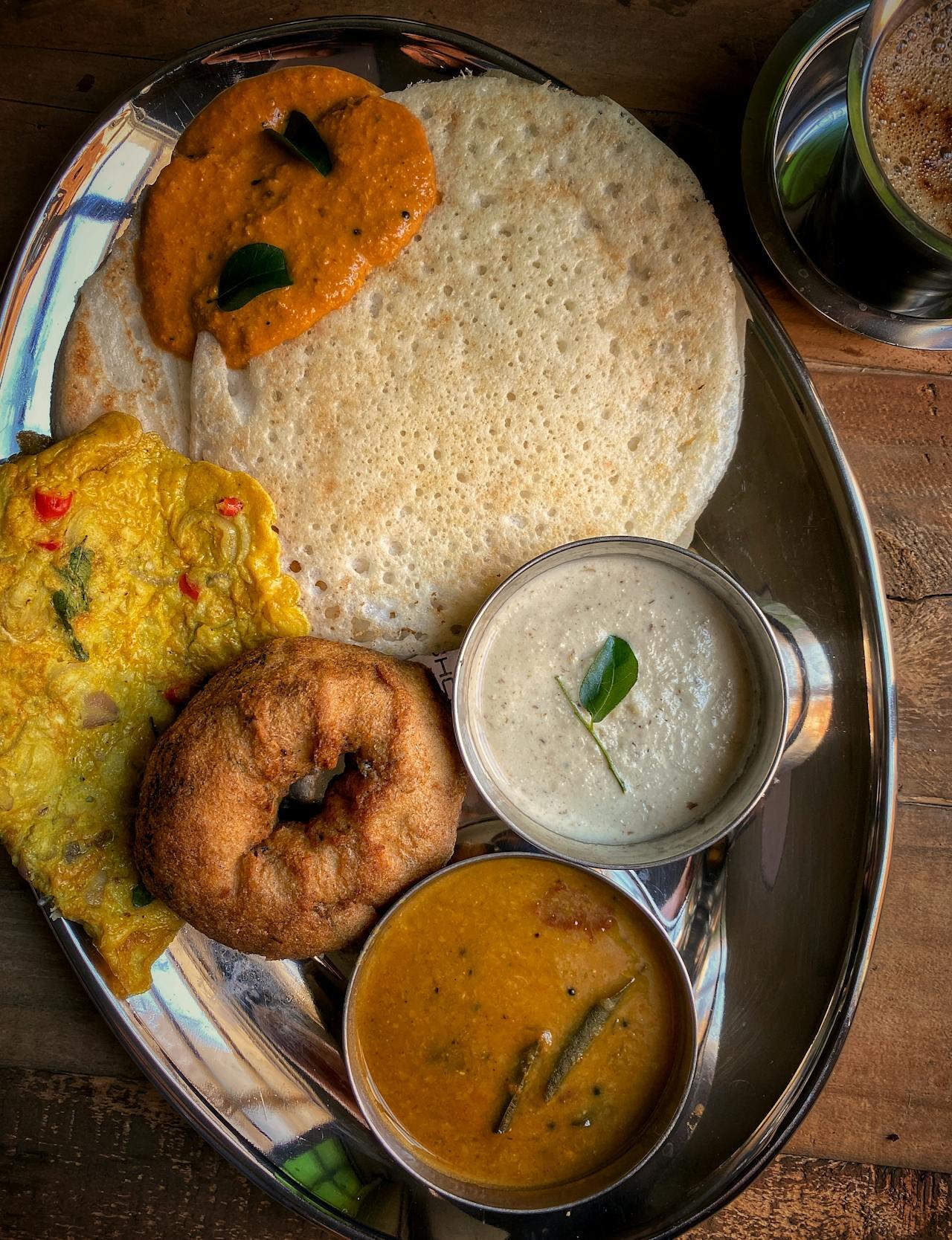 A stainless steel platter featuring crispy dosa, a soft idli, and a donut-shaped vada, accompanied by three colorful chutneys and sambar.