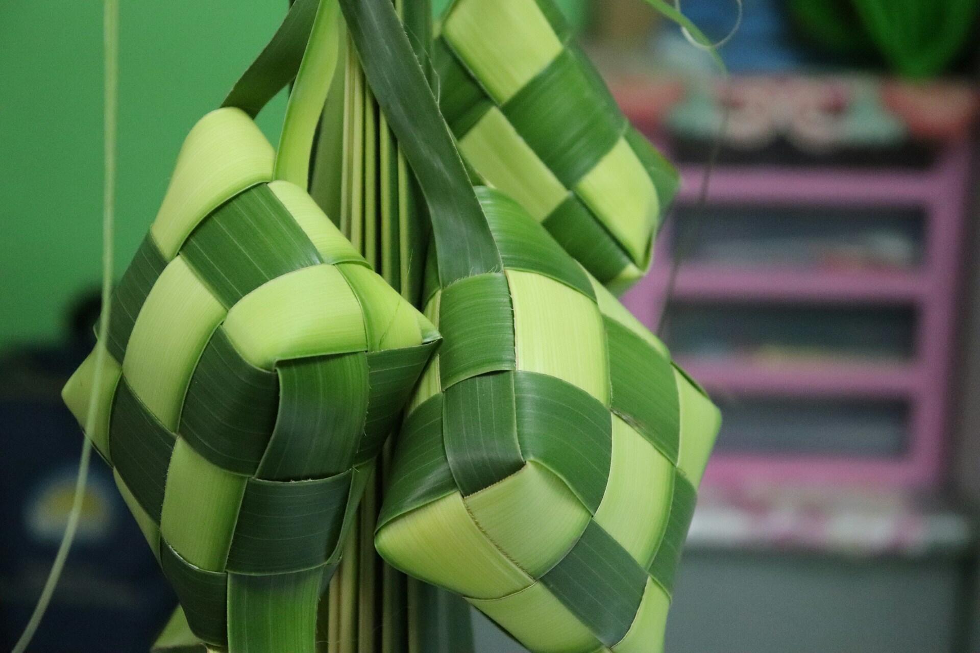 Three intricately woven, green and yellow rice dumplings (ketupat) hung against a soft green backdrop, showcasing traditional craftsmanship.