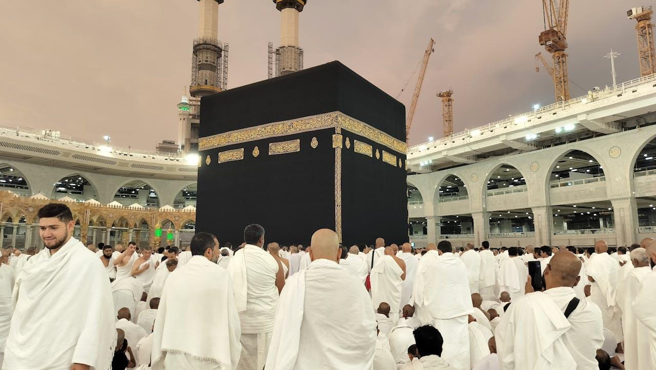 A large crowd of pilgrims in white clothing surrounds the Kaaba, a black cube structure, during a spiritual gathering in Mecca.