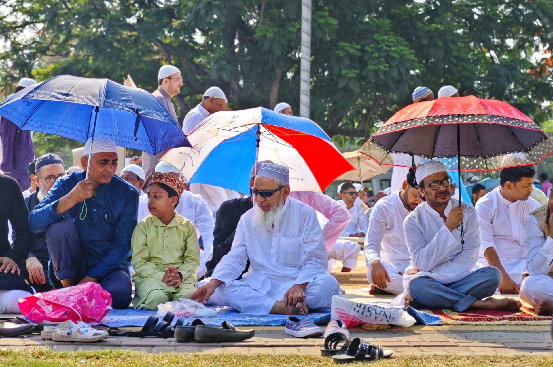 A group of people seated on the ground under colorful umbrellas, preparing for a prayer gathering in an outdoor setting.