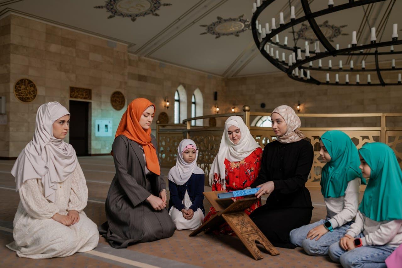 A group of women and girls in hijabs is gathered around a wooden book holder, engaged in a learning session inside a spacious mosque.