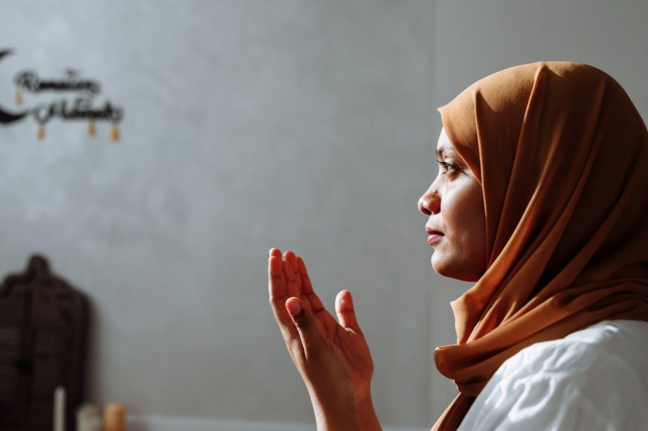 A woman in a brown hijab and white attire is seen in profile, with her hands raised in prayer against a softly lit background.