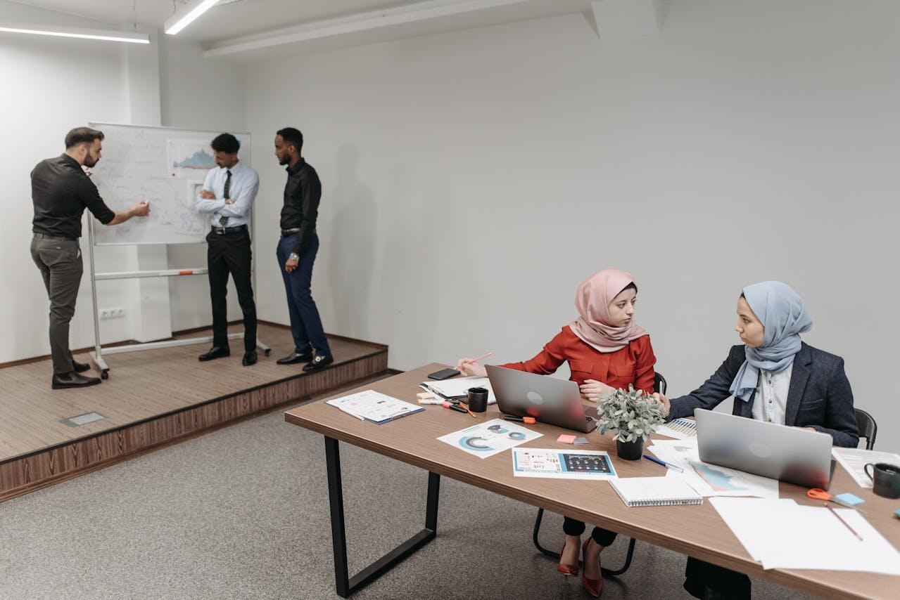 A modern office meeting scene with professionals discussing at a whiteboard and others working on laptops at a table.