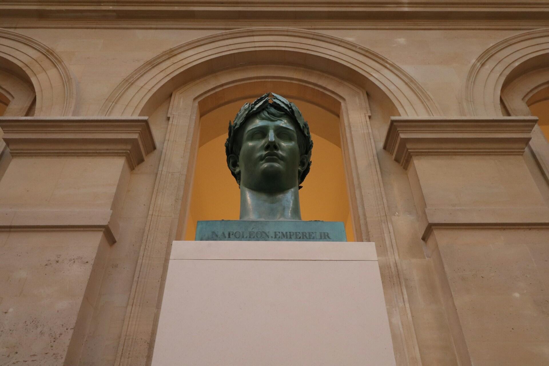 A bronze bust of Napoleon Bonaparte displayed within an elegant architectural setting, featuring arched walls and soft lighting.
