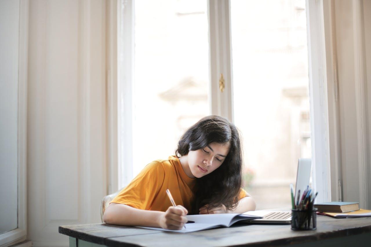 A person in an orange shirt writes in a notebook at a desk with a laptop and colorful pens, sitting near bright windows.