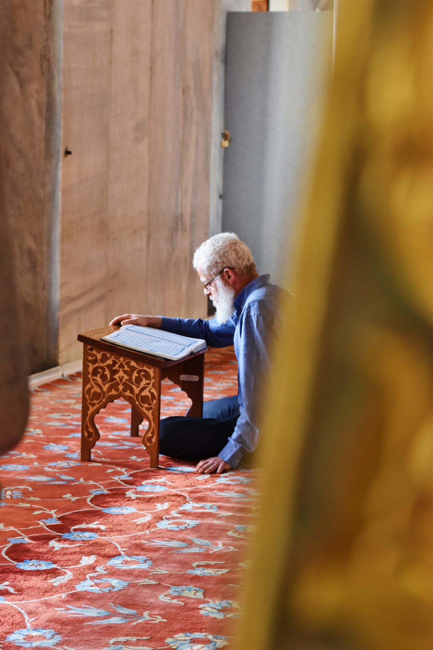 A person sits cross-legged on an ornate rug, studying a book placed on a small wooden stand in a tranquil indoor setting.