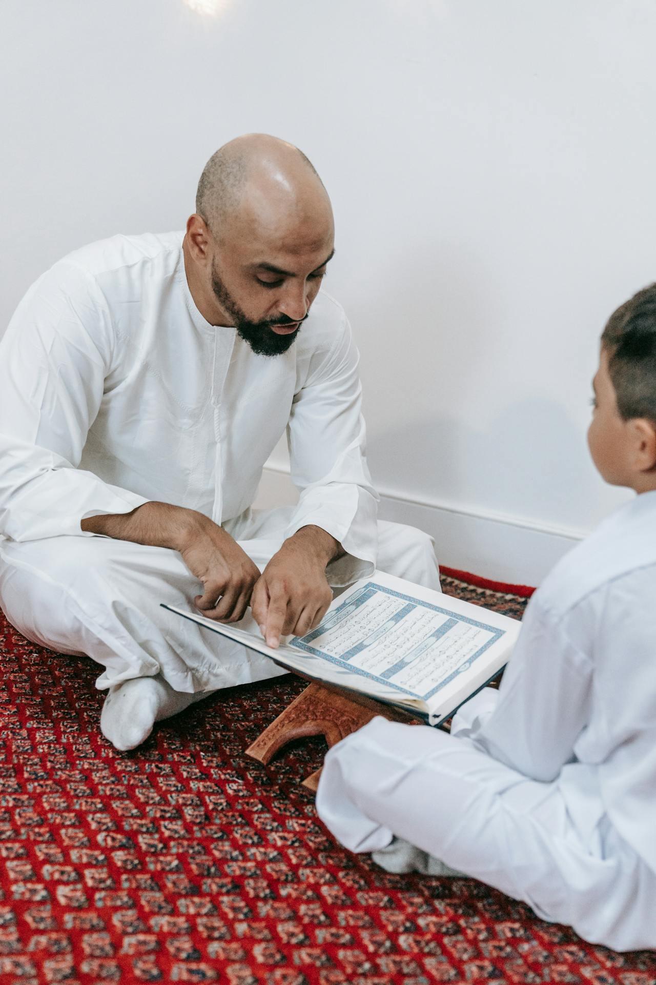 An adult in traditional white attire teaches a child, referencing a book, on a patterned red carpet in a bright room.