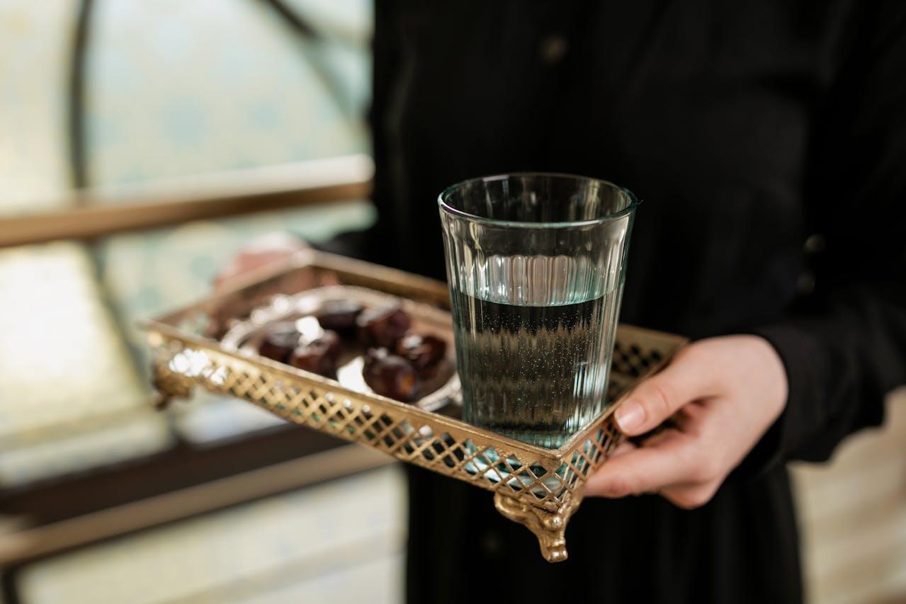 A person in a black outfit holds a decorative tray with a glass of water and a plate of dates.