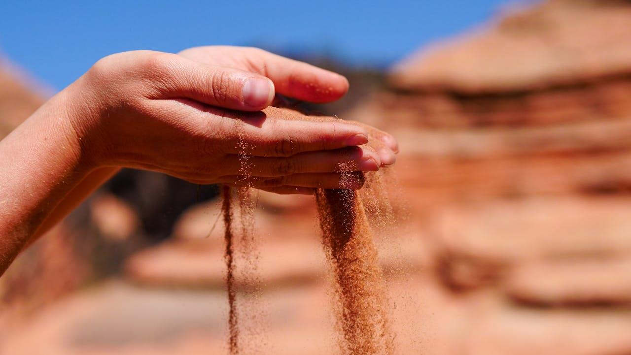 A hand gently pours sand between its fingers against a blurred, warm-toned landscape and a clear blue sky.