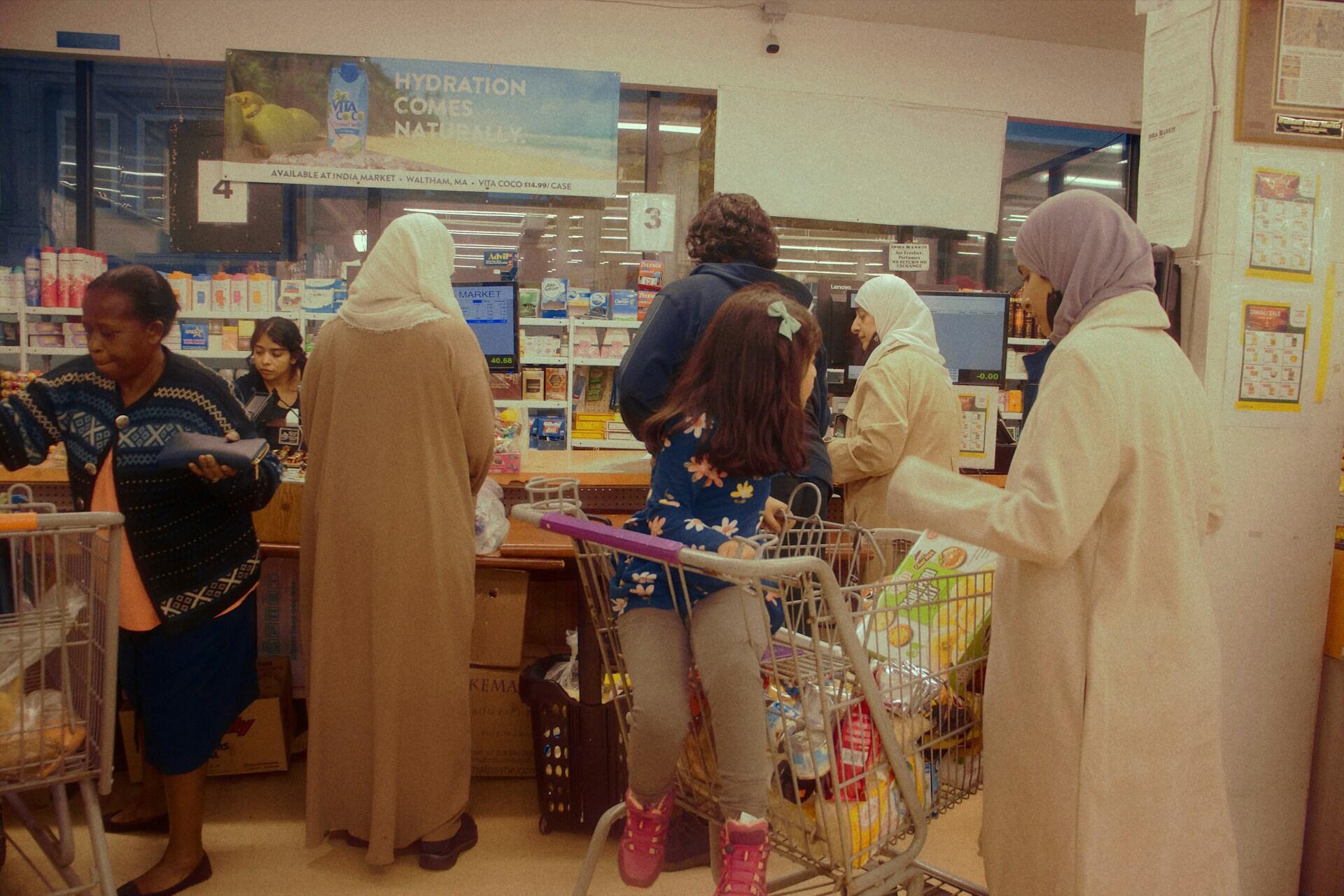 A group of people at a grocery store checkout, with shopping carts full of items and a sign that reads "Hydration Comes Naturally."