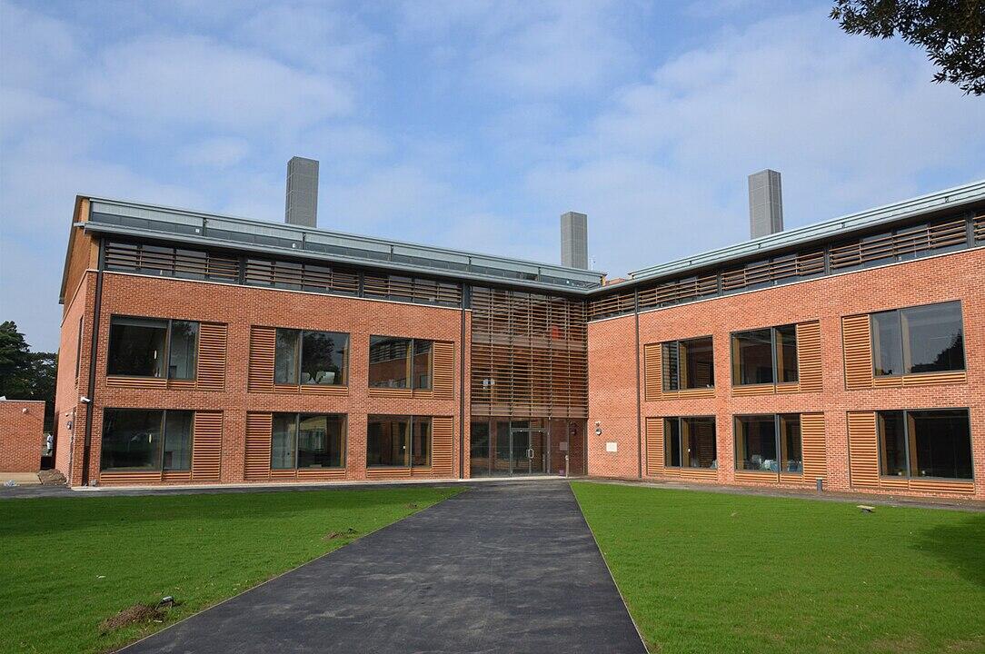 Modern red brick building with large windows and wooden slats, featuring a landscaped lawn and clear blue sky.