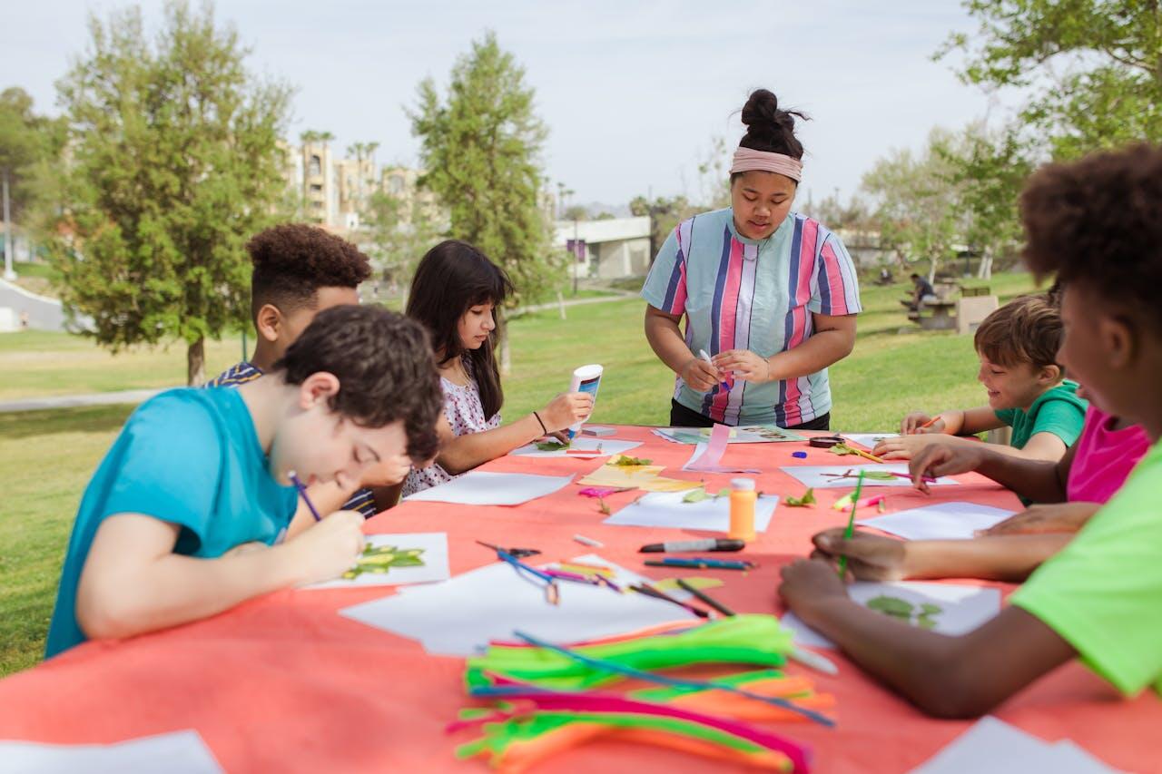 A diverse group of children engaged in an arts and crafts activity outdoors, surrounded by colorful materials and a bright green park.