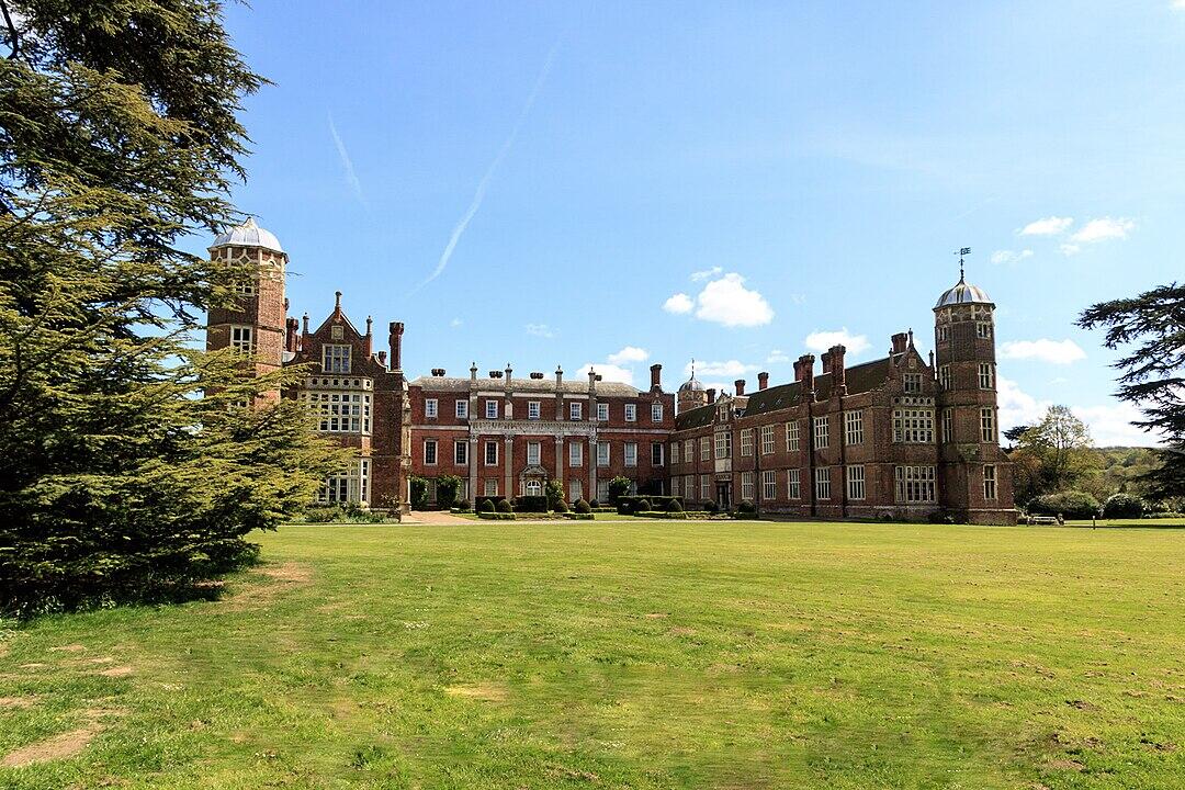 A grand, historical mansion with a mix of architectural styles, surrounded by well-maintained lawns and clear blue skies.