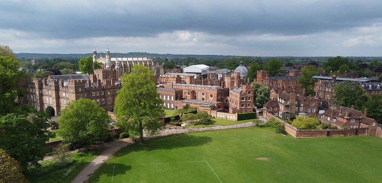 Aerial view of a grand, historic campus with red brick buildings, lush greenery, and a cloudy sky in the background.