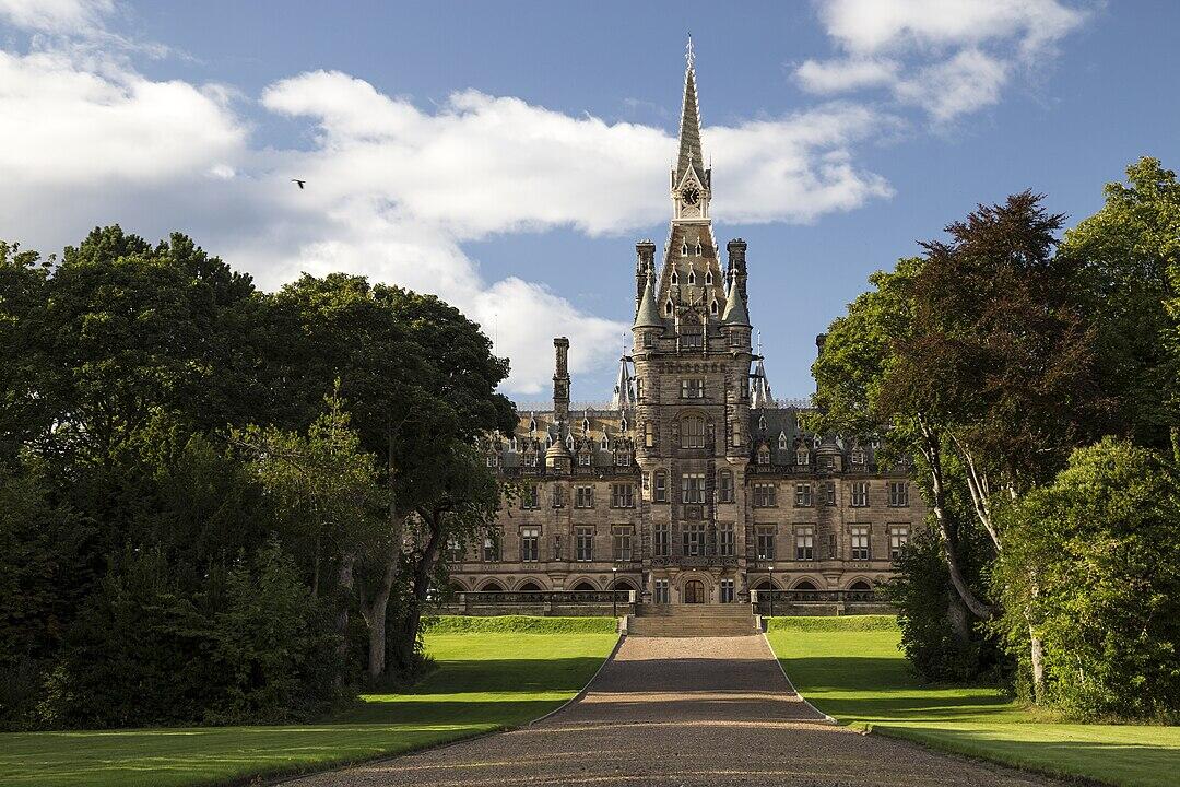 A grand castle with a tall spire, surrounded by lush greenery and blue skies, featuring a wide pathway leading to its entrance.