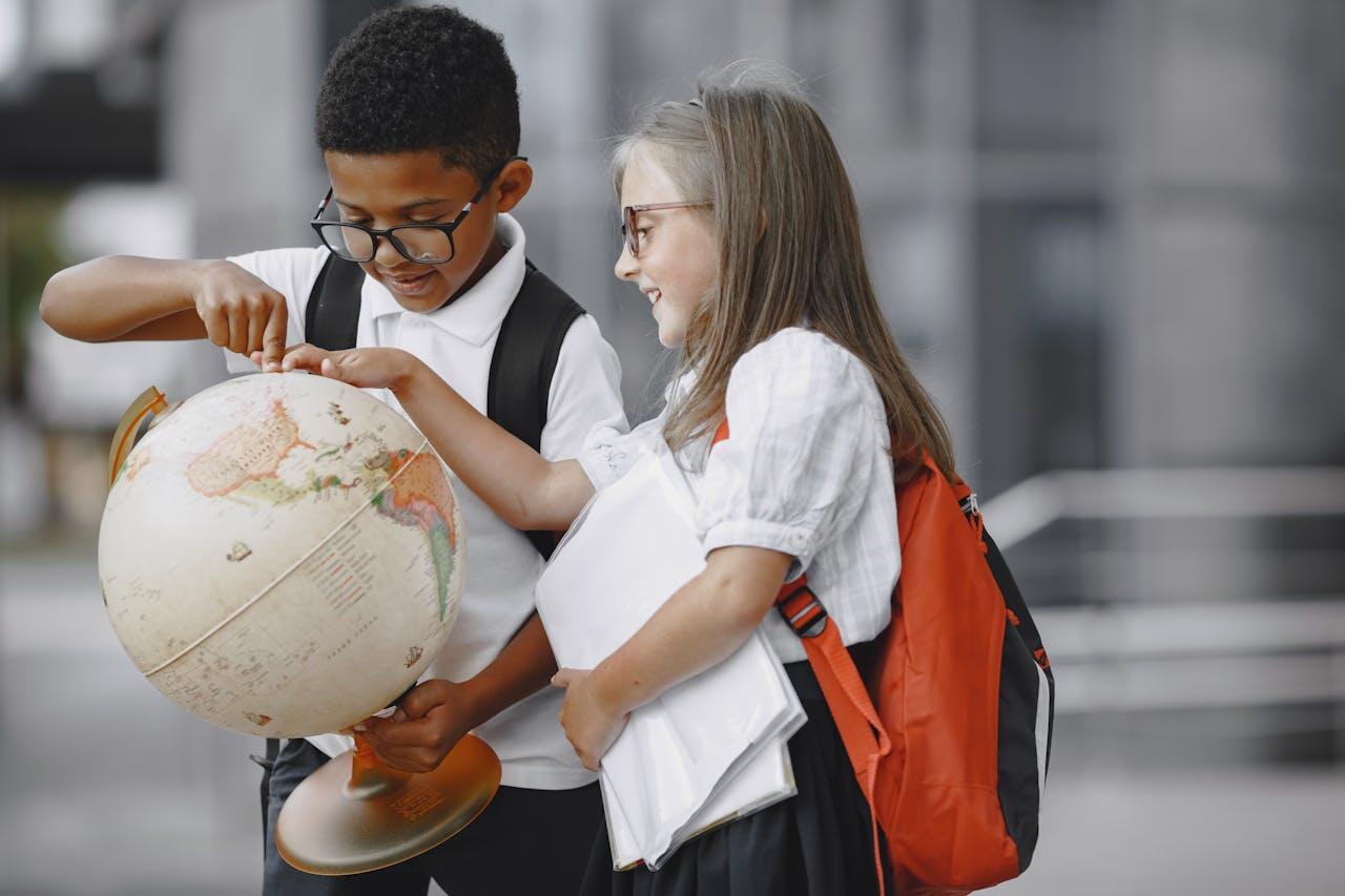 Two children explore a globe outdoors, engaged in a discussion. One has a black backpack, and the other carries school materials.